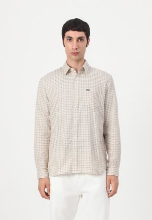 Uomo con camicia beige a quadri abbottonata a maniche lunghe e pantaloni bianchi, in piedi davanti a uno sfondo chiaro e semplice.