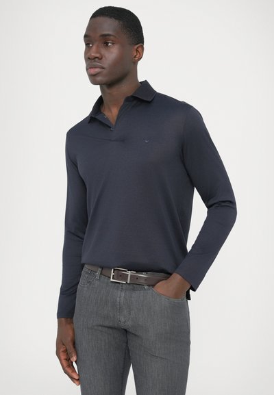 Tmavomodrá polokošeľa s dlhým rukávom a golierom, s malým logom na hrudi, doplnená sivými textúrovanými nohavicami a hnedým opaskom.