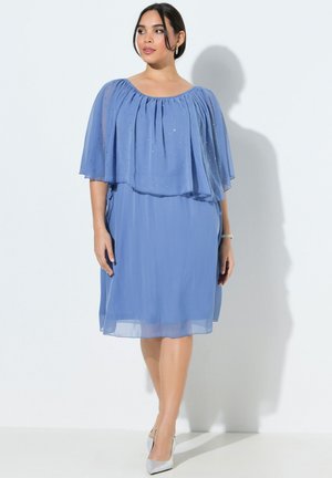 Femme portant une robe bleue en chiffon longueur genou avec une cape transparente ornée et des talons hauts argentés, debout contre un mur blanc.