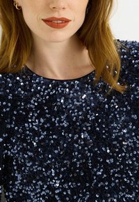 Top azul marino con lentejuelas y cuello redondo, con una superficie texturizada de lentejuelas superpuestas que crean un efecto brillante.