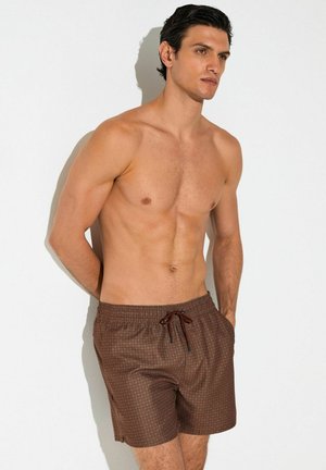 Homme torse nu aux cheveux foncés portant un short de bain marron à motifs, debout, les mains derrière le dos, contre un mur blanc.