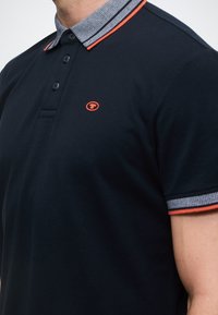 Meripaitapusero, jossa on teksturoitu kangas, ja siinä on erottuva kaulus punaisilla yksityiskohdilla sekä pieni logo rinnan kohdalla. Lyhyet hihat.