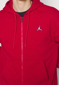 Felpa rossa con zip realizzata in cotone, dotata di una tasca a marsupio, cordini regolabili e un logo in bianco e nero sul petto.