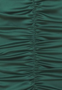 Smaragdgrön satin tyg med samlad textur som löper vertikalt. Den släta ytan framhäver glansen och skapar en mjuk, draperad effekt.