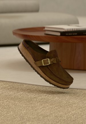 Brauner Slipper aus Wildleder mit einer Korksohle, verziert mit einer dekorativen Metallschnalle und einer Schleifenakzentuierung, vor einem hellen, strukturierten Hintergrund.