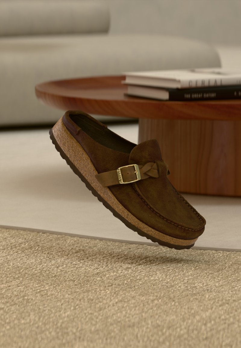 Καφέ γκρίζο παπούτσι με σούστες και σόλα από φελλό, διακοσμημένο με μεταλλικό αγκράφα και φιόγκο, τοποθετημένο πάνω σε ανοιχτό υφασμάτινο φόντο.