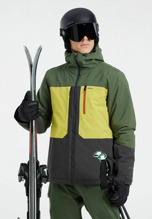 Chaqueta de esquí verde y amarilla con una sección inferior negra, equipada con un bolsillo con cremallera, acompañada de guantes negros y esquís plateados.