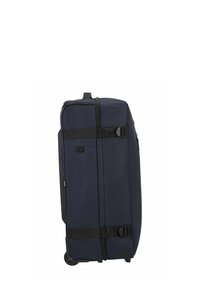Valise à roulettes en tissu bleu marine, avec une surface texturée, des sangles noires et deux roues pour la mobilité. Poche latérale zippée visible.