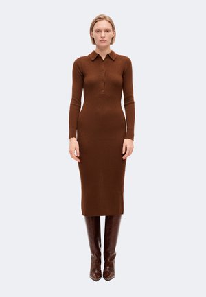Femme portant une robe midi marron moulante à manches longues côtelées, avec un col et des boutons, associée à des bottes hautes marron foncé.