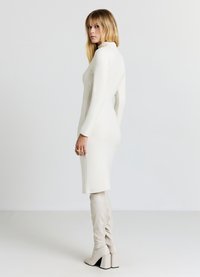 Figurbetontes cremefarbenes Kleid mit langen Ärmeln, hohem Kragen und knielangem Saum. Kombiniert mit kniehohen beigen Stiefeln und goldenen Ringen als Akzente.