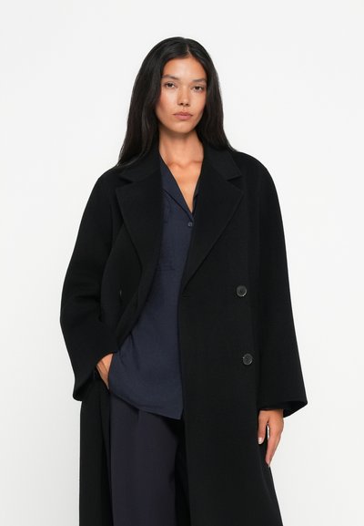 Schwarzer Wollmantel mit Reverskragen, doppelt geknöpft und weiten Ärmeln. Kombiniert mit einem marineblauen Hemd darunter. Minimalistisches Design.
