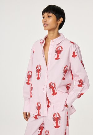 Person trägt lockeres pinkfarbenes Hemd und Hose mit rotem Hummerdruck, steht mit einer Hand in der Tasche und schaut leicht zur Seite.