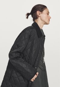 Giacca nera trapuntata con colletto in corduroy, dotata di bottoni a pressione e una superficie liscia, indossata su un top grigio. Vista laterale.