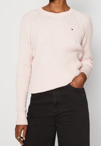 Vaaleanpunainen teksturoitu neulepusero, jossa on pitkät hihat ja ribbireunus kaula-aukossa. Puserossa on pieni, monivärinen logo rinnassa.