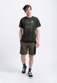 Donkergroen t-shirt met een graphic van bergen en cactussen, gecombineerd met olijfgroene cargo shorts met zijzakken en zwarte sneakers met witte zolen.
