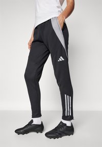 Black athletic pants with gray side panels, featuring zippered cuffs and three white stripes on the lower leg, made from a smooth fabric.