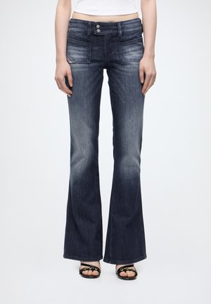 Kvinde iført mørkeblå vide jeans med forlommer og dobbelt knaplukning, kombineret med sorte sandaletter med åben tå.