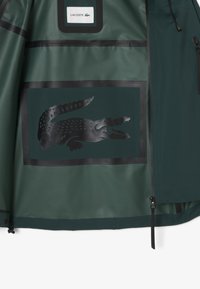 Groene waterdichte jas interieur met een groot reliëf zwart krokodillenlogo met een rechthoekige rand en een Lacoste-label erboven.