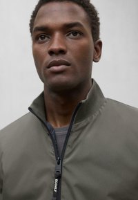 Chaqueta con cremallera de color verde oliva y cuello alto, fabricada en un tejido suave; cuenta con una cremallera negra con la marca "ECOALF".