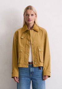Afgekorte gele jas met een corduroy kraag, voorzien van drukknoopsluitingen, voorzakken en een rits. Combinatie met lichtblauwe spijkerbroek.