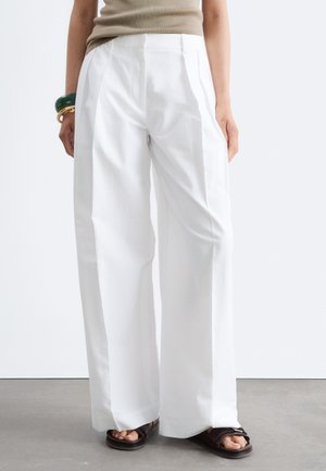 Personne portant un pantalon blanc à jambes larges, un haut beige sans manches, des sandales foncées et des bracelets verts et dorés au poignet gauche.