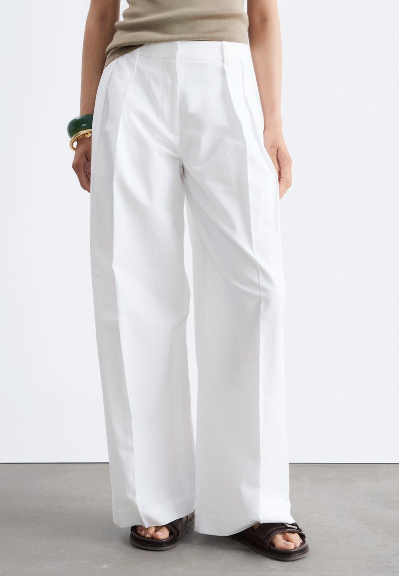 Personne portant un pantalon blanc à jambes larges, un haut beige sans manches, des sandales foncées et des bracelets verts et dorés au poignet gauche.