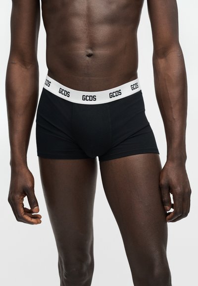 Ανδρικός κορμός που φοράει μαύρα αντρικά εσώρουχα τύπου μποξεράκια με λευκή ζώνη μέσης, διακοσμημένη με επαναλαμβανόμενο λογότυπο "GCDS", στέκεται μπροστά σε απλό ανοιχτό φόντο.