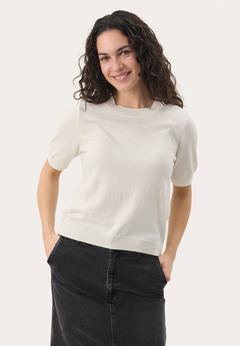 Pull léger beige à manches courtes avec un col rond et un ourlet côtelé, associé à une jupe en denim noire. Texture douce, design décontracté.