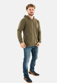 Haut de survêtement zippé vert olive avec capuche et poches latérales, porté avec un jean bleu et des baskets noires avec des accents orange.