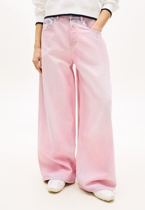 Osoba ubrana w jasnoróżowe szerokie spodnie jeansowe, białe trampki i biały długi rękaw, z jedną ręką w kieszeni.