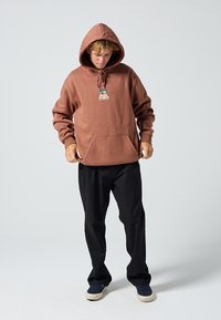 Smeđa dukserica s kapuljačom i prednjim džepom, s grafičkom oznakom. Nošena s crnim hlačama i mornarsko plavim cipelama na neutralnoj pozadini.