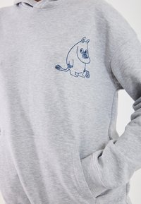 Šedá mikina s ilustrací Moomin postavy v námořnické modři na levém prsu, s klokankou a měkkou texturou.