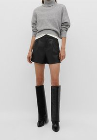 Suéter gris de punto con cuello alto, combinado con pantalones cortos negros de cuero plisados. La modelo lleva botas altas de cuero negras con puntera puntiaguda.