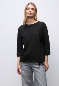 Lyshåret kvinde iført en sort, langærmet top med en lille sløjfedetalje og grå, vidde bukser, stående mod en ensfarvet, lys baggrund.