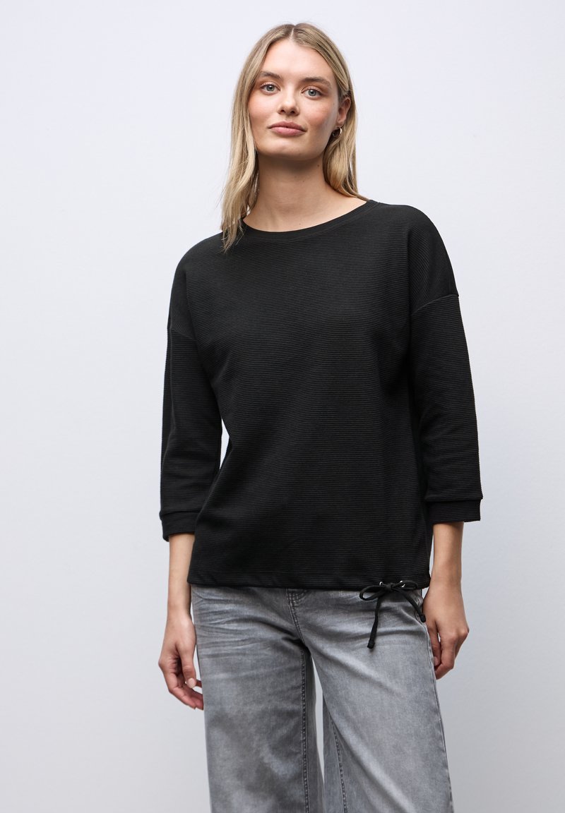 Lyshåret kvinde iført en sort, langærmet top med en lille sløjfedetalje og grå, vidde bukser, stående mod en ensfarvet, lys baggrund.