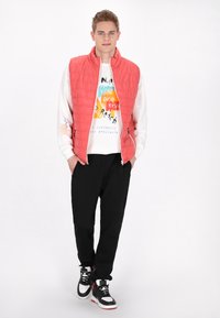 Gilet trapuntato corallo sopra una felpa bianca con grafica, abbinato a pantaloni neri e sneaker neri con accenti rossi. In piedi su uno sfondo bianco.
