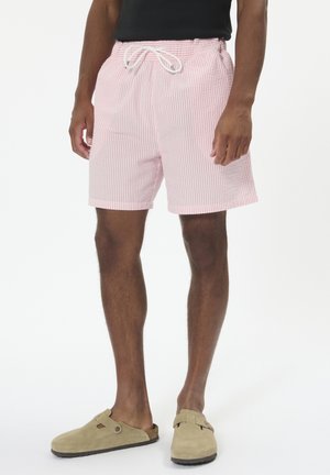 Gestreifte rosa-weiße Badehose mit elastischem Bund und Kordelzug. Das Material wirkt leicht; getragen mit beigen Slipper aus Wildleder.