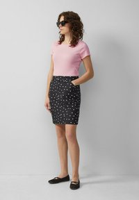 Frau mit lockigem Haar, die schwarze Sonnenbrille, ein pinkes Kurzarmoberteil, einen schwarzen Rock mit weißen Punkten und schwarze Ballerinas trägt und steht.