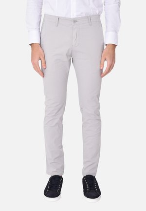 Uomo che indossa pantaloni slim fit grigio chiaro, camicia bianca elegante e sneakers nere, in piedi dritto su sfondo bianco.