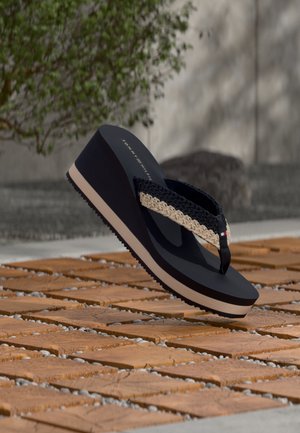 Tongs noires à plateforme avec des brides tissées beige et noire, posées sur un sol en carrelage terracotta à l'extérieur.