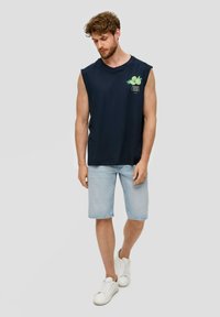 Donkerblauw mouwloos T-shirt met ronde hals, met een groene grafische print. Gemaakt met lichtblauwe denim shorts en witte sneakers.