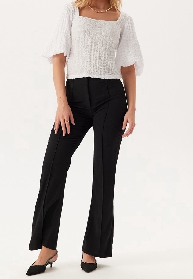 Haut blanc texturé avec manches courtes bouffantes, associé à un pantalon noir ajusté et des talons noirs à bouts pointus. Design simple et élégant.