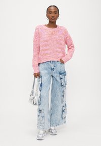 Rozā un oranžs adīts džemperis ar apaļu kakla izgriezumu, kombinēts ar gaiši ziliem plaša piegriezuma cargo džinsiem ar kabatām un sudraba krāsas sporta apaviem.