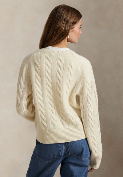 Femeie purtând un pulover crem cu model împletit și mâneci lungi și blugi albaștri, văzută din profil pe un fundal simplu.