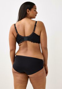 Reggiseno in pizzo nero con schiena a rete, dotato di spalline regolabili e bordo ondulato. Abbinato a slip neri senza cuciture, che mettono in evidenza una texture liscia.