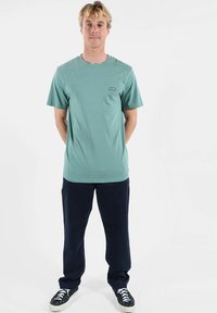 T-shirt en coton vert clair avec un col rond, des manches courtes, et un petit logo sur la poitrine gauche. Porté avec un pantalon bleu marine foncé et des chaussures noires.