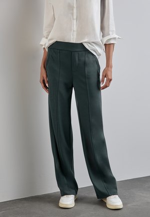 Pantalons larges verts avec des coutures verticales, en tissu lisse, associés à une chemise blanche à boutons et des baskets blanches.