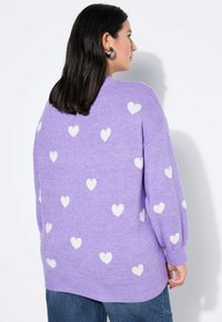 Fioletowy sweter wykonany z miękkiego materiału, zdobiony wzorem białych serc. Luźny krój z prążkowanymi mankietami i dolnym brzegiem. Noszony na niebieskich dżinsach.
