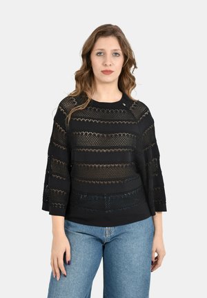 Donna con capelli mossi indossa una maglia nera a manica lunga con motivo in pizzo e jeans azzurri chiari, in piedi davanti a uno sfondo grigio chiaro uniforme.