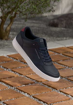 Black and red Tommy Hilfiger sneaker with white sole resting above wooden square tiles and small stones outdoors.